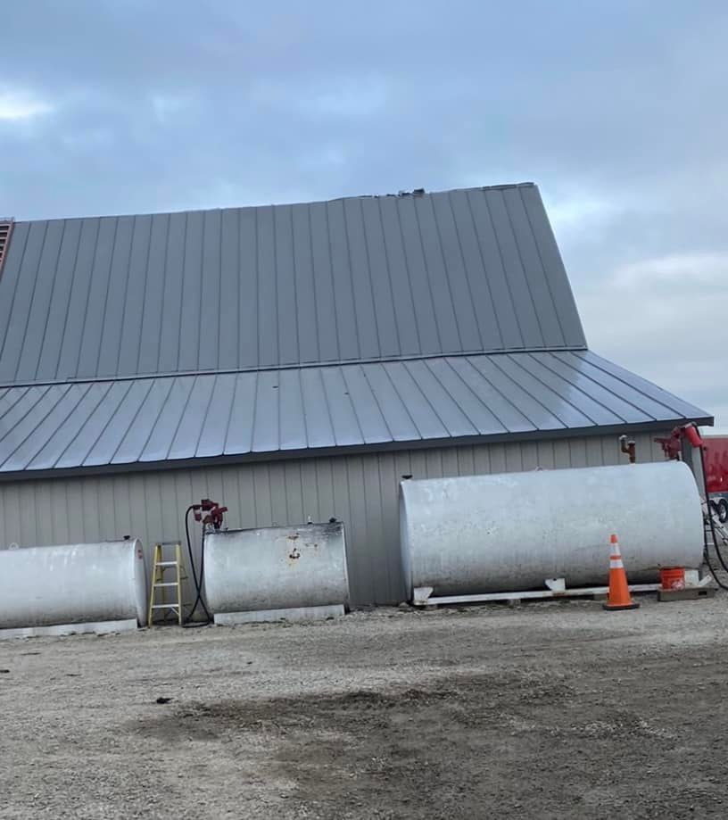 Large gray metal building with two white tanks, ladders, and orange cones on a gravel lot under cloudy sky