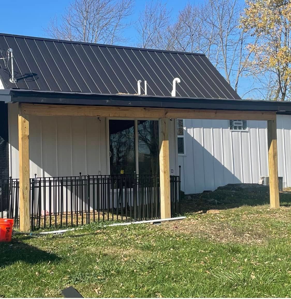 Small white house with a black metal roof, wooden porch posts, and black railing in a grassy yard under blue sky