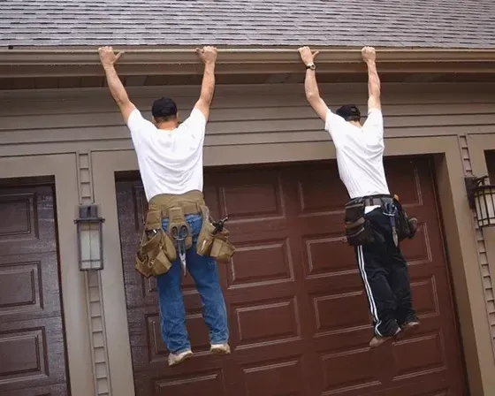 Two workers in white shirts hanging from a house roof edge above a garage door