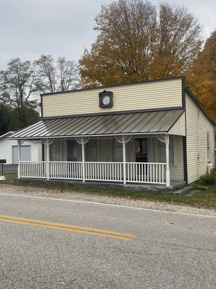 Small beige storefront with a metal awning and white porch railing beside a road.