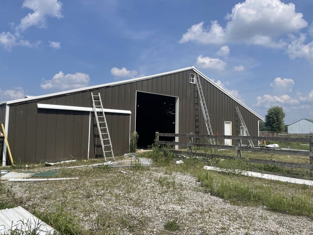 Large gray barn with open doors and ladders outside on a gravel lot under a cloudy sky