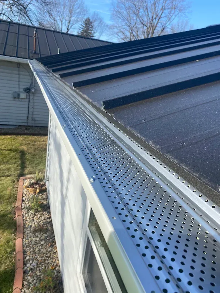 Perforated metal gutter along a dark sloped roof beside a house on a sunny day