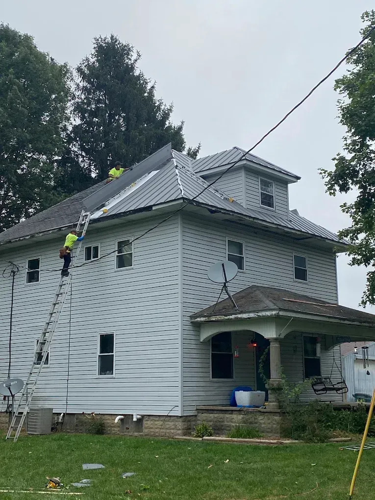 Worker on ladder repairing the gray roof of a white house under cloudy sky