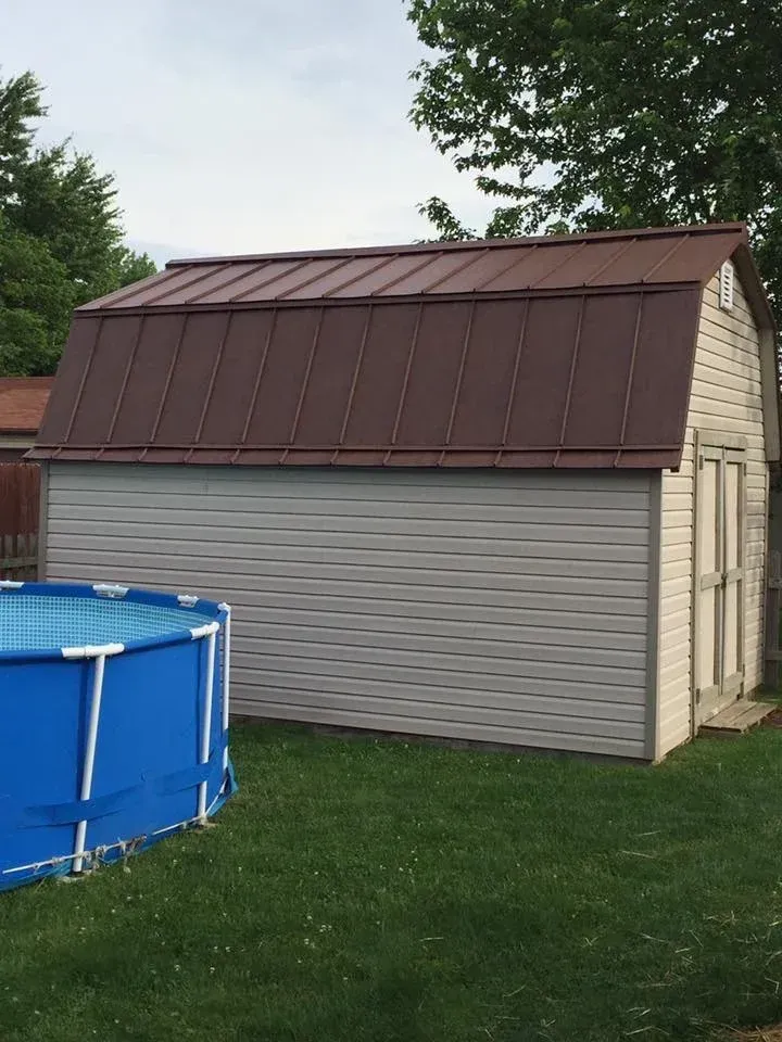 Small shed with a brown metal roof and gray siding beside a blue above-ground pool on a grassy yard.