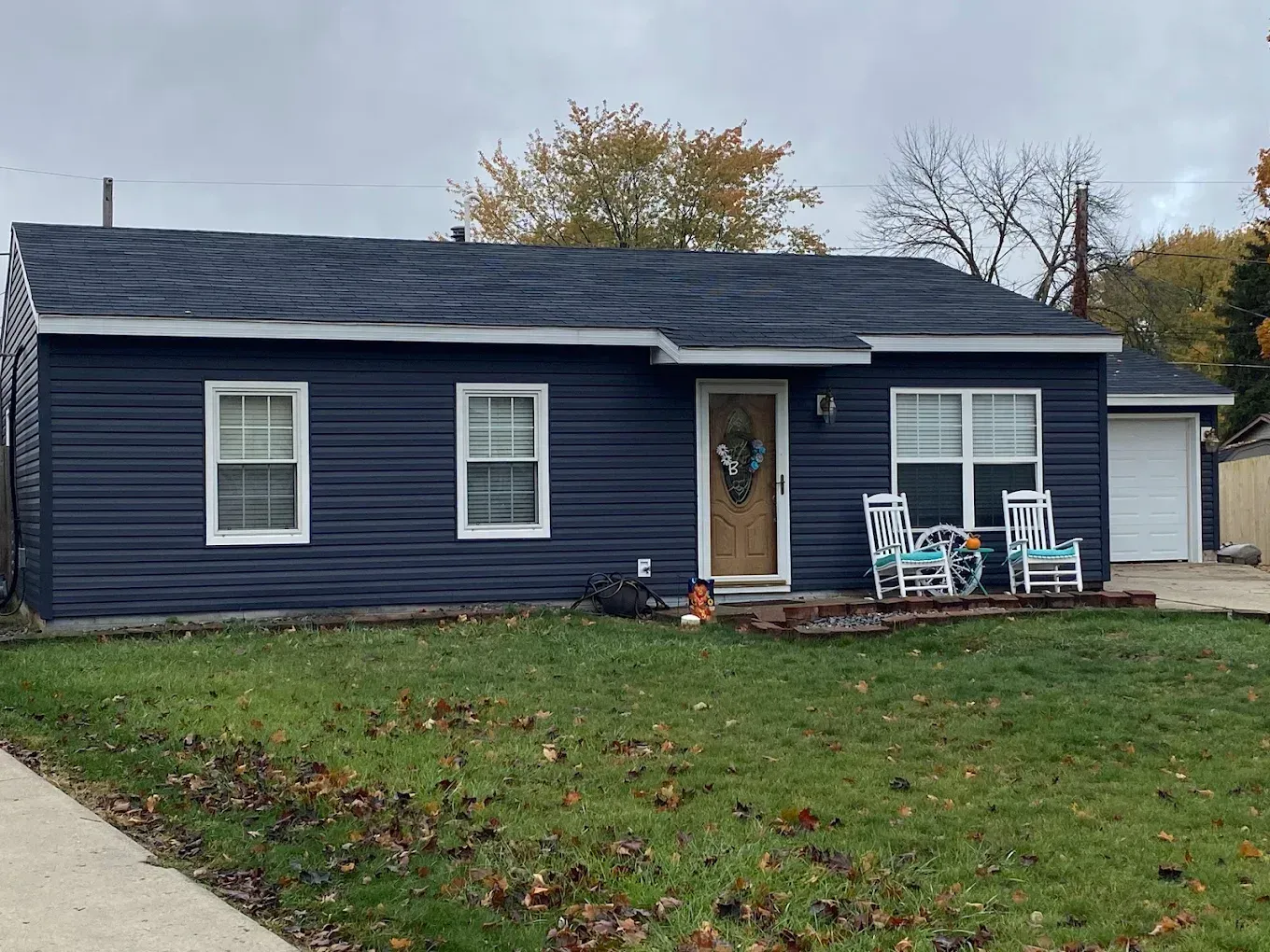 Single-story dark blue house with white trim, front porch chairs, and a small lawn in overcast weather