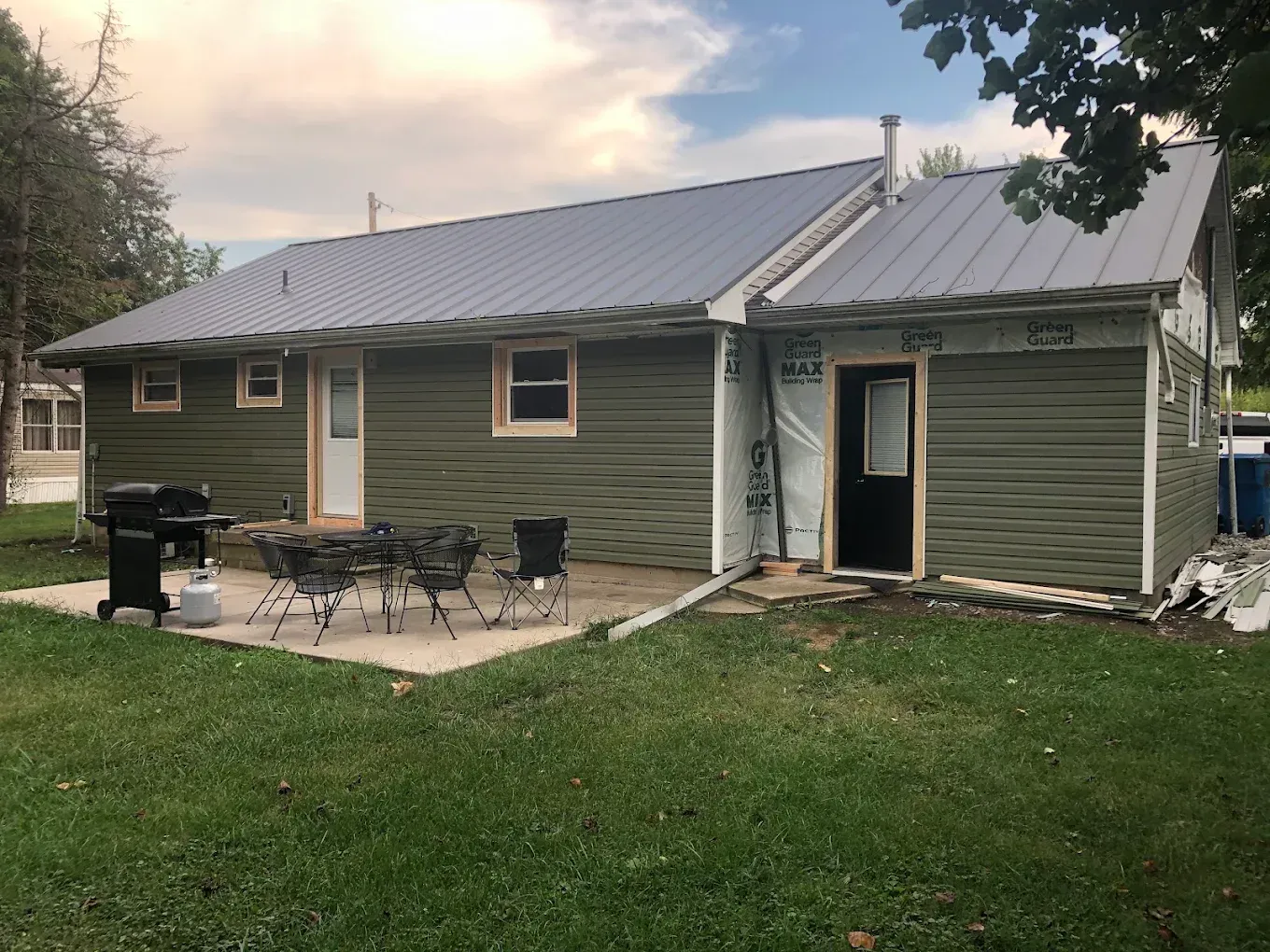 Green house with a metal roof, patio furniture, grill, and open side door in a grassy yard at dusk