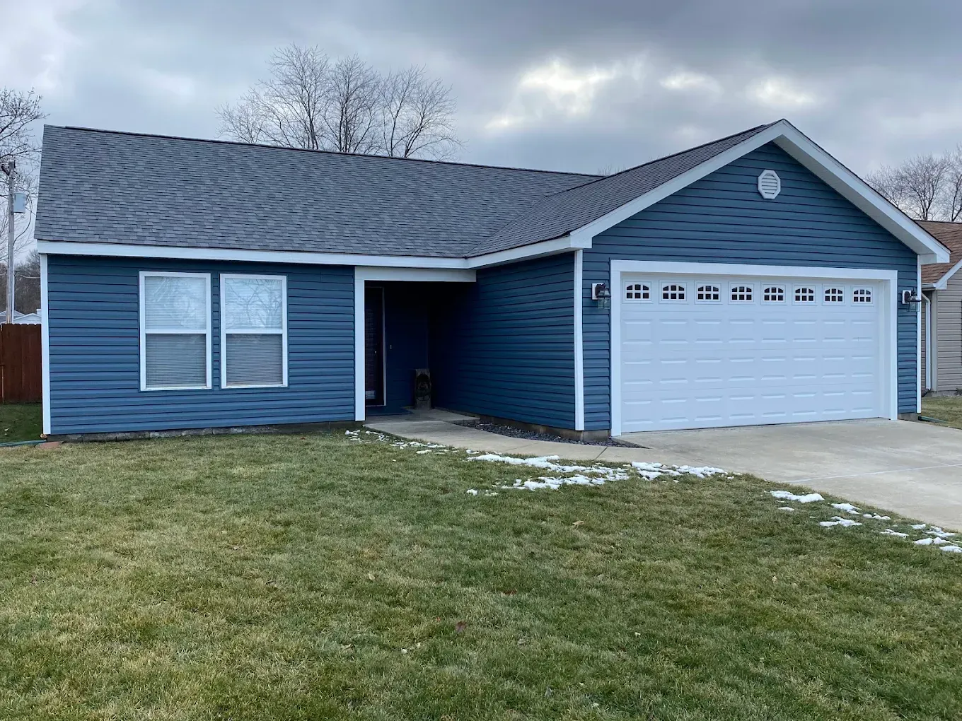 Blue single-story house with attached garage and patchy lawn under a cloudy sky