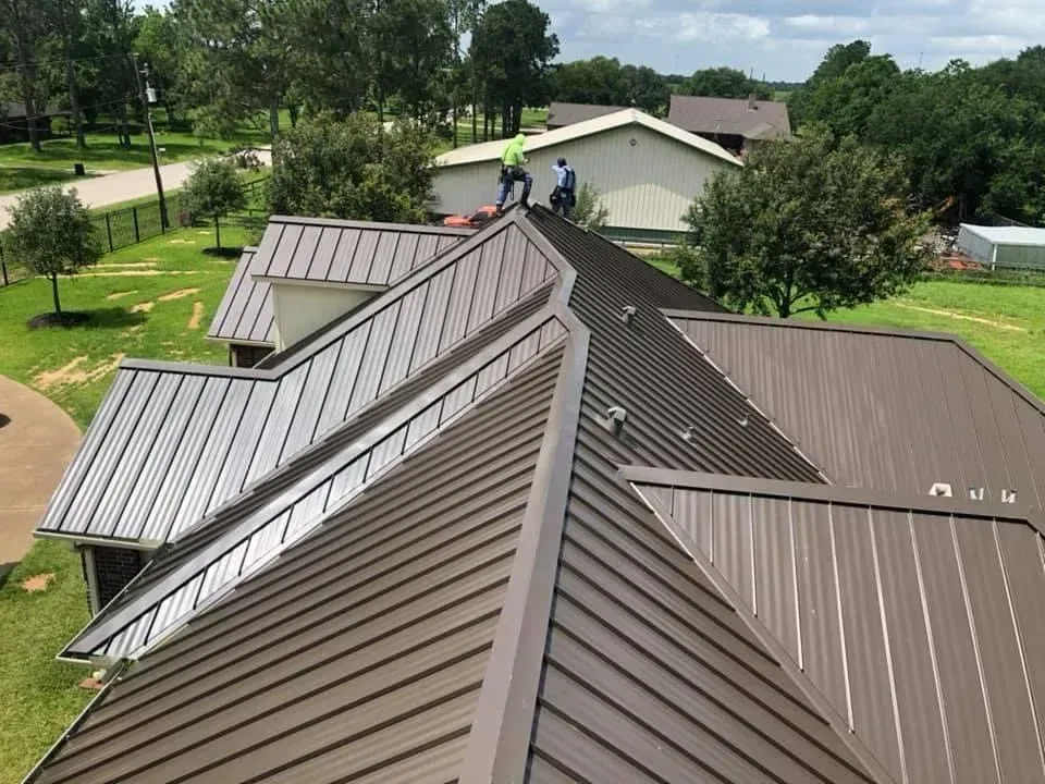 Roof with metal panels and solar panels, viewed from above near a tree-lined residential area.