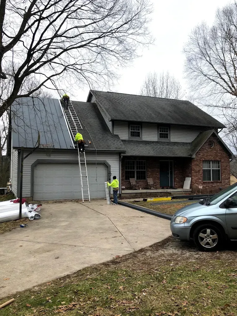 Workers on ladders repairing a house roof and garage in a suburban driveway.