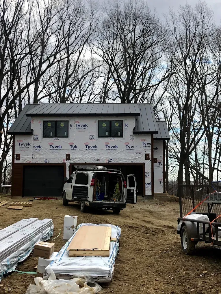 Partially built house with wraparound scaffolding, construction truck, and lumber piles in a wooded yard