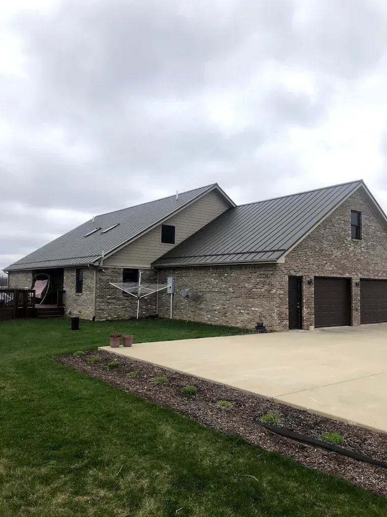 Stone house with gray metal roof, garage doors, and a green lawn under an overcast sky