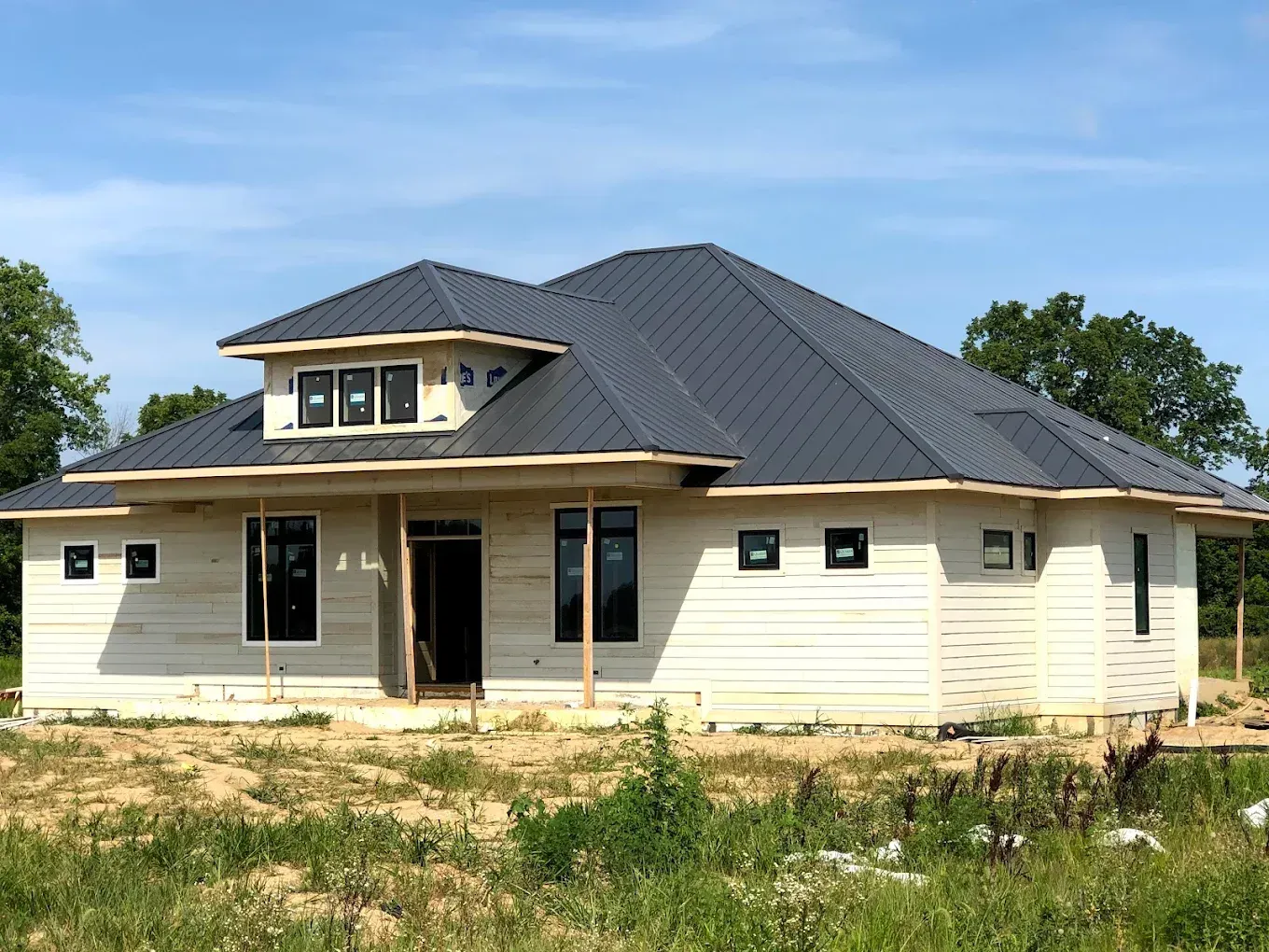 New house under construction with a dark gray roof and cream siding, surrounded by bare ground and trees.