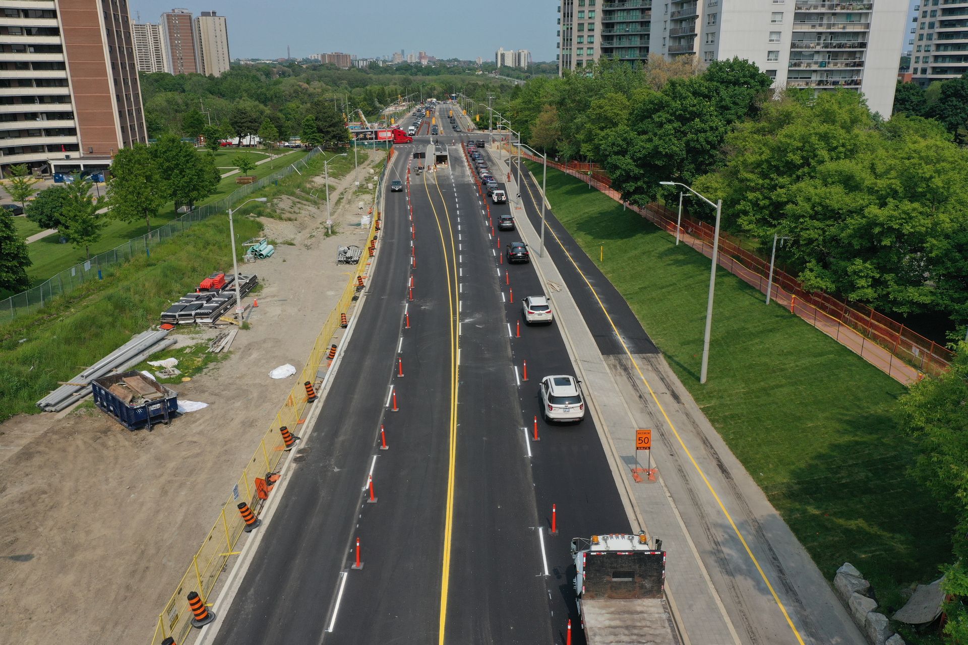 Road under construction with cars driving on newly paved sections. Buildings and trees visible.