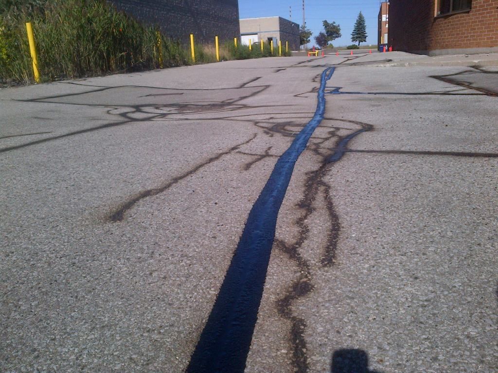 Asphalt road with a dark sealant down the middle and cracks. Buildings and vegetation in the background.