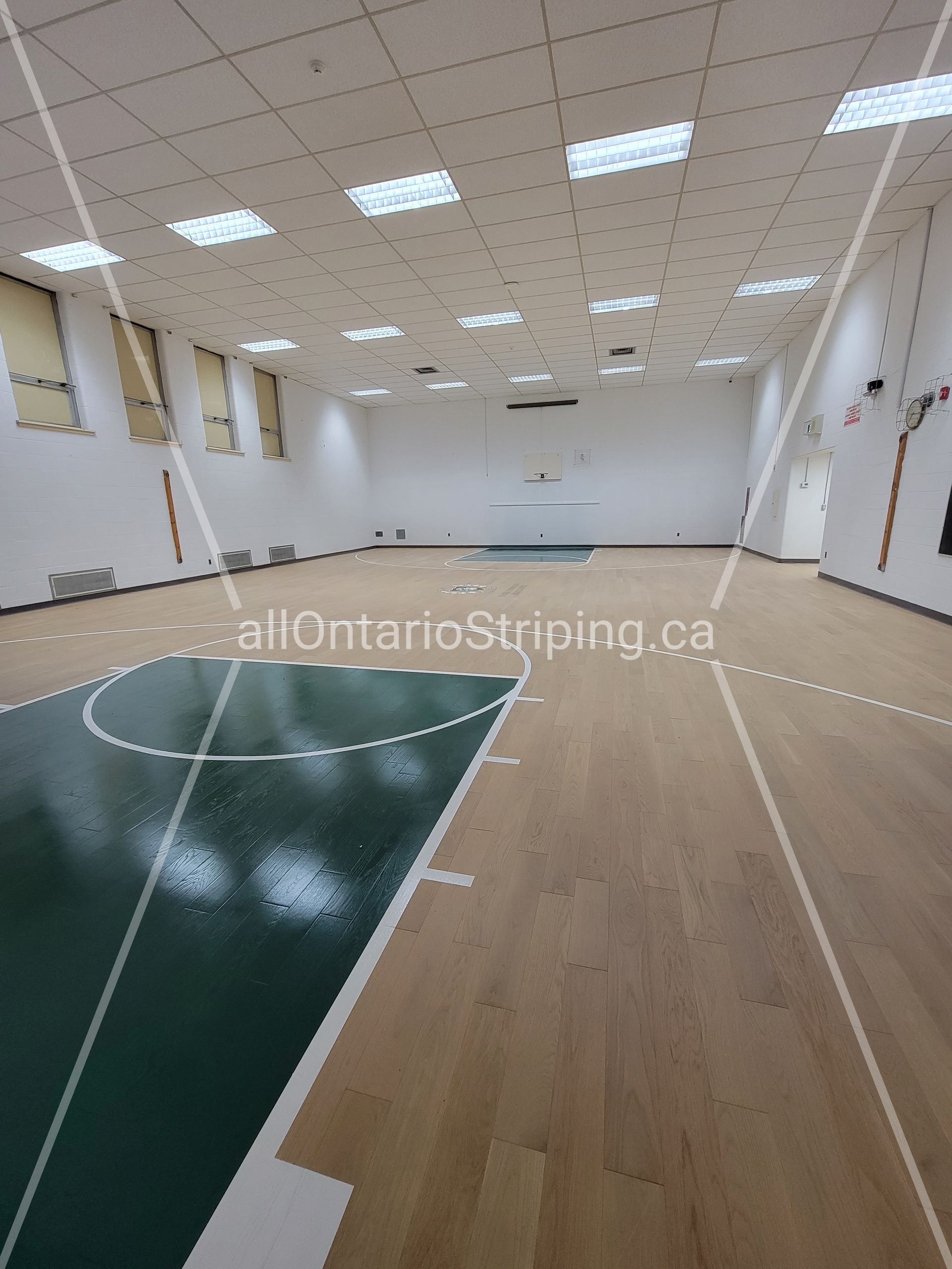 Empty gymnasium with hardwood floor, green basketball court markings, and bright overhead lighting.