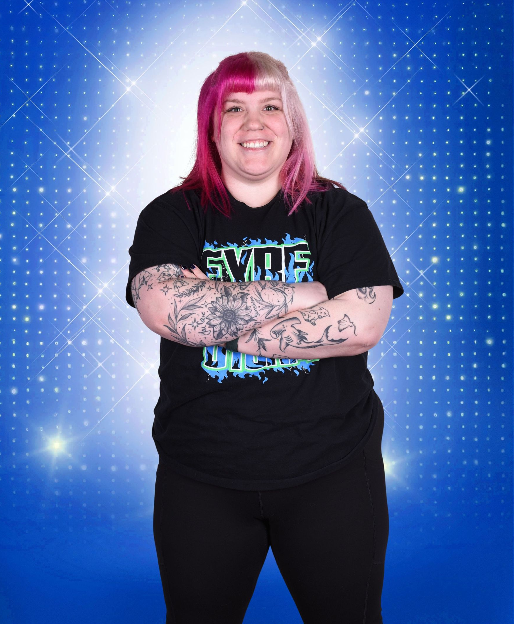 Person with pink hair and tattooed arms standing against a blue starry backdrop, arms crossed.