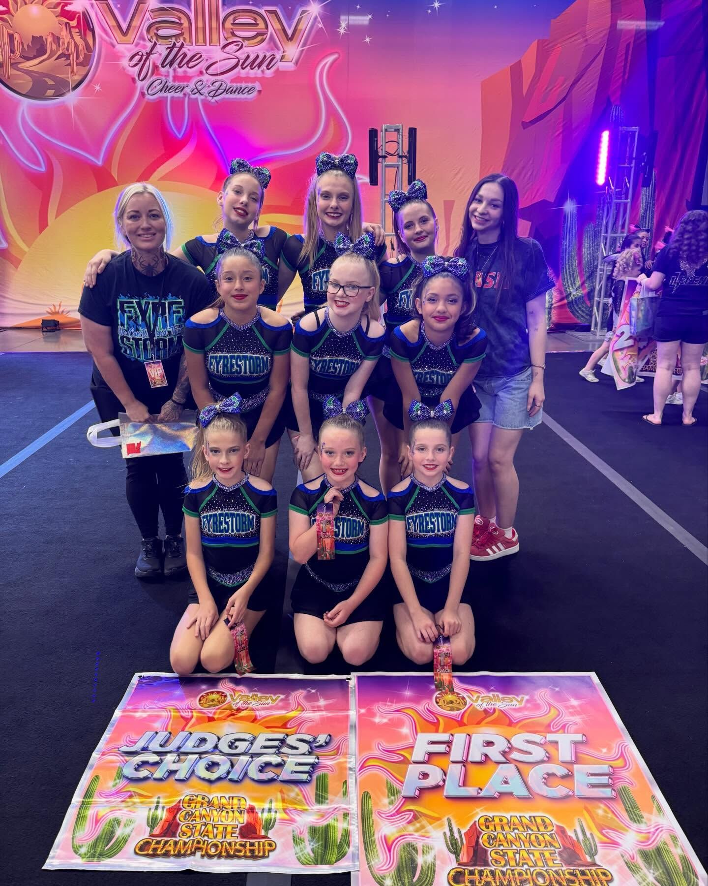 Cheer team posing on a competition floor with two large first-place signs, under pink and purple lights