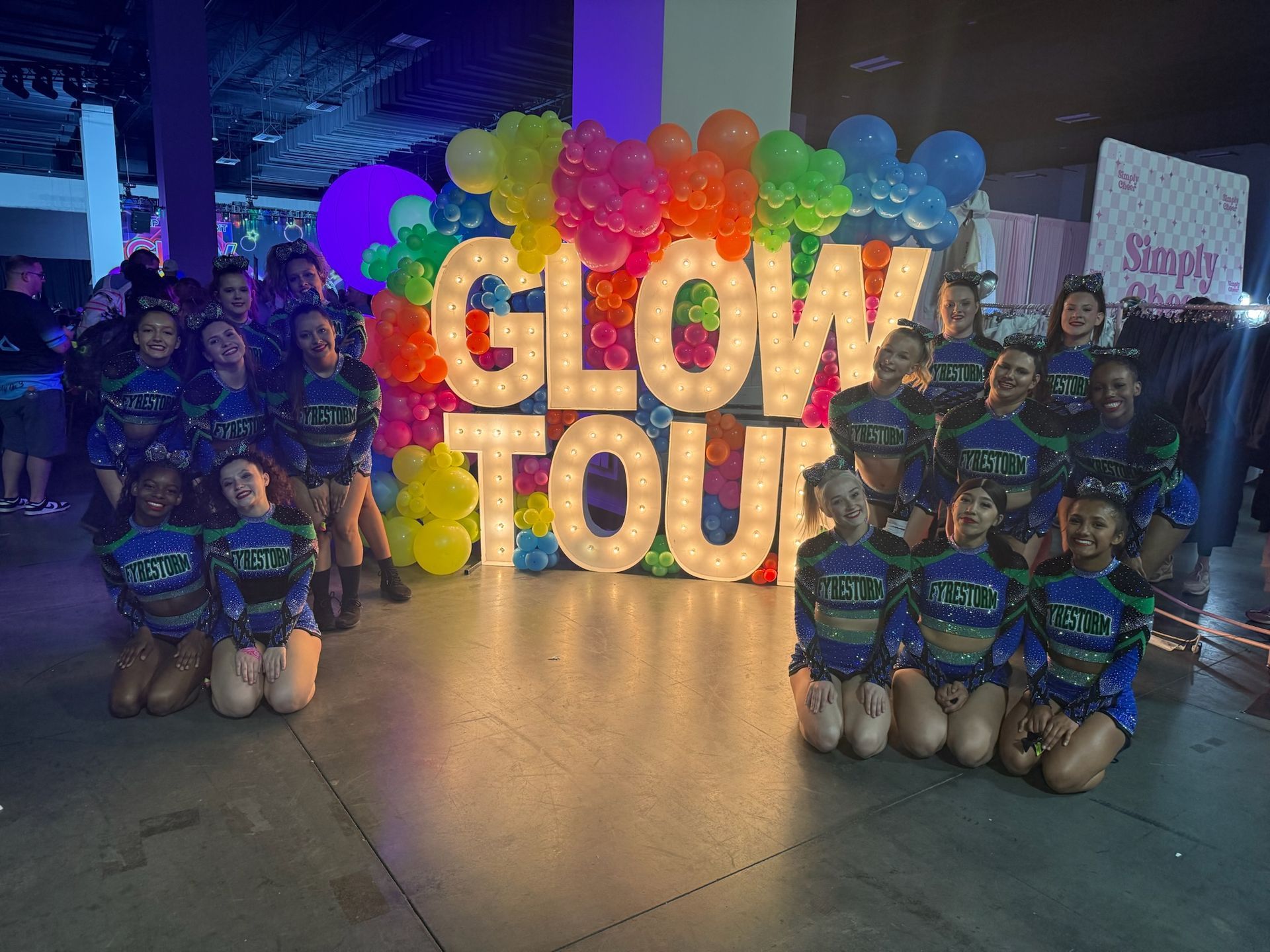 Group of children in blue outfits posing around a colorful “GLOW TOUR” balloon display at night