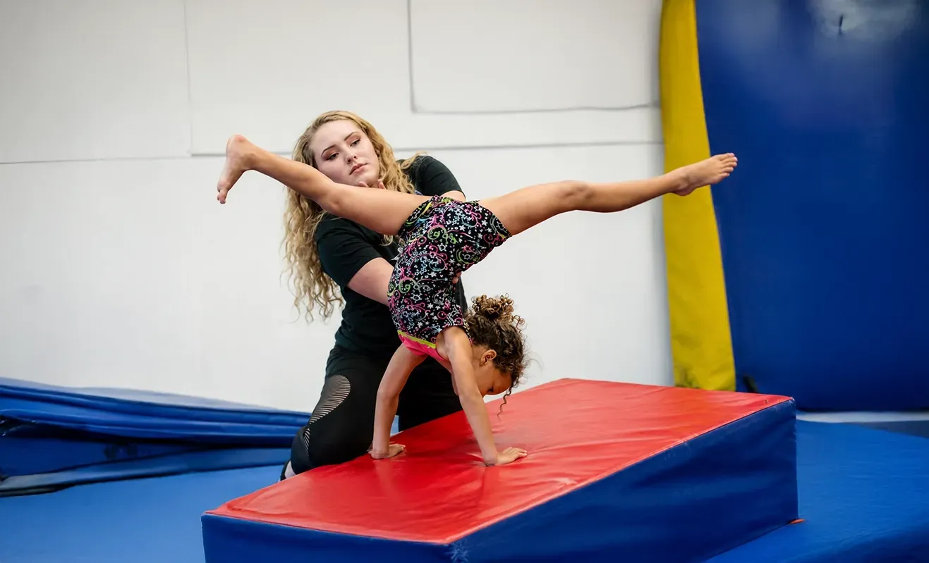 Gymnast balancing on a mat with arms and leg extended, with a coach spotting her in a gym