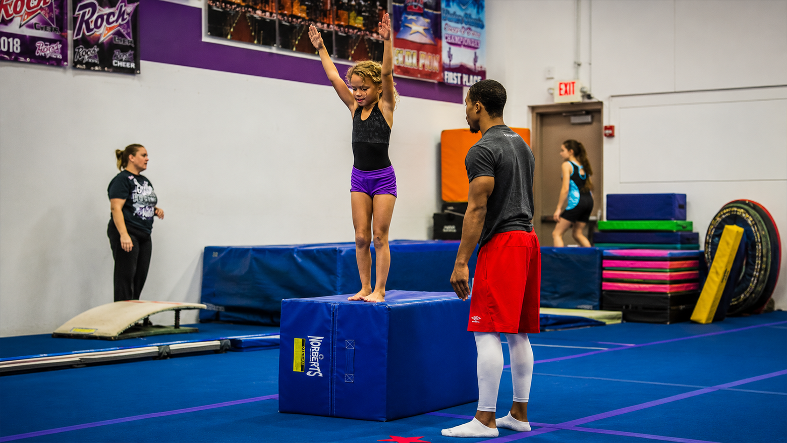 Gymnastics class with a girl balancing on a blue foam block while others watch in a training gym