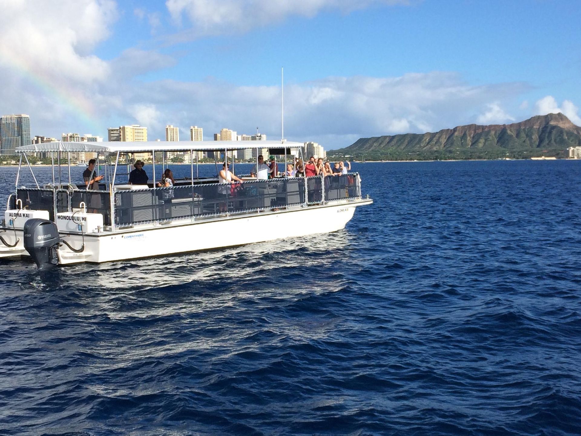 Tour boat with passengers on blue water near a coastal skyline and rocky island under a partly cloudy sky