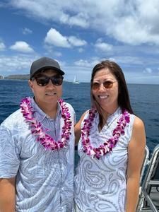 Two people wearing pink leis pose on a boat with blue ocean and cloudy sky behind them.