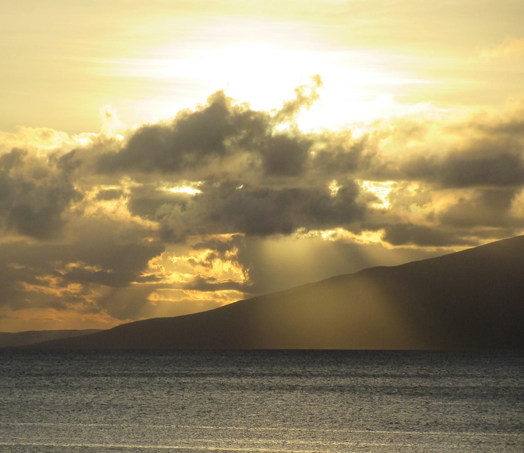 Golden sunset over the ocean with dark clouds and sun rays behind a coastal hillside