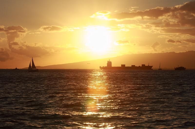 Sunset over the ocean with sailboats and a cargo ship silhouetted on the horizon