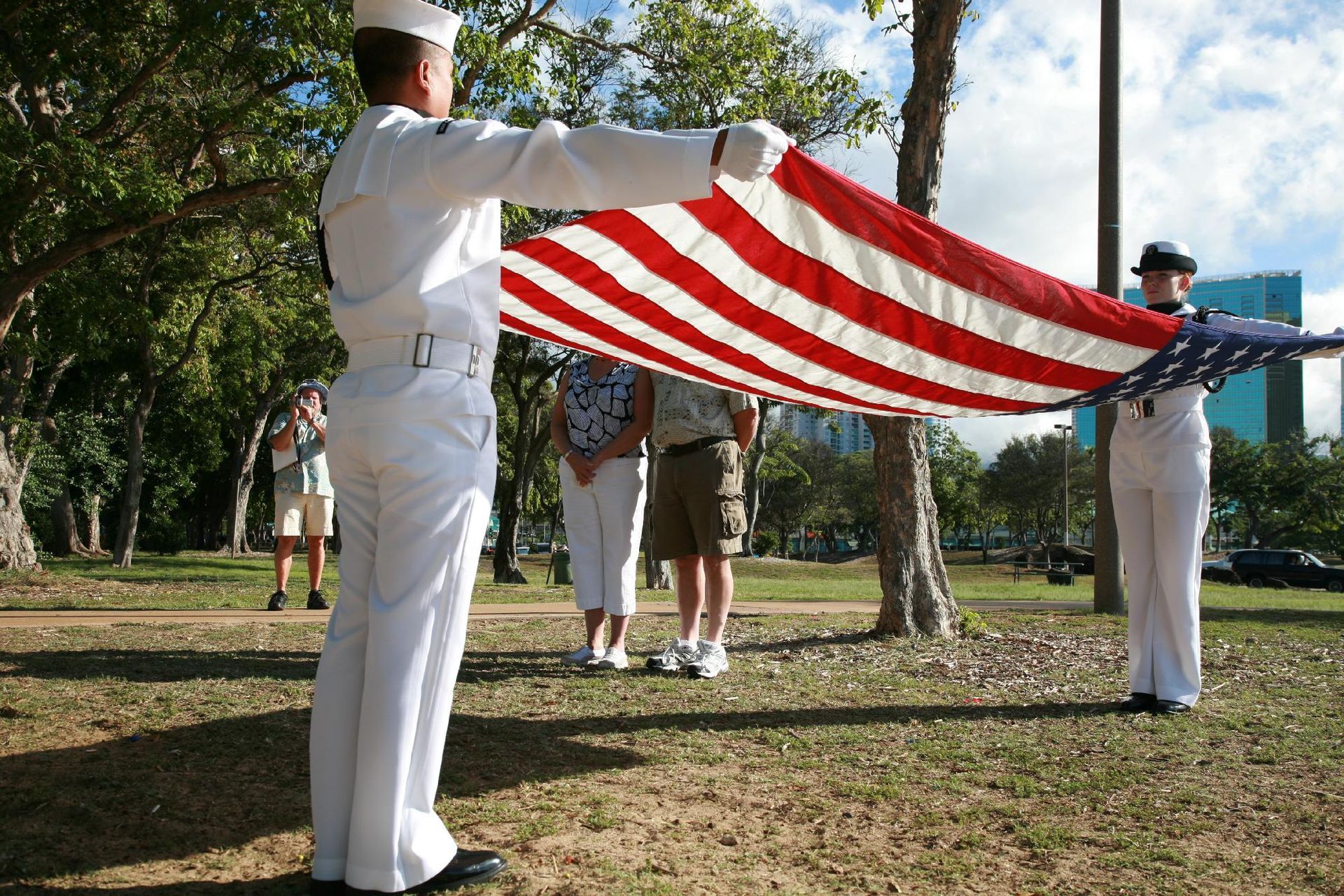 People in white uniforms holding and folding a large American flag outdoors in a park.