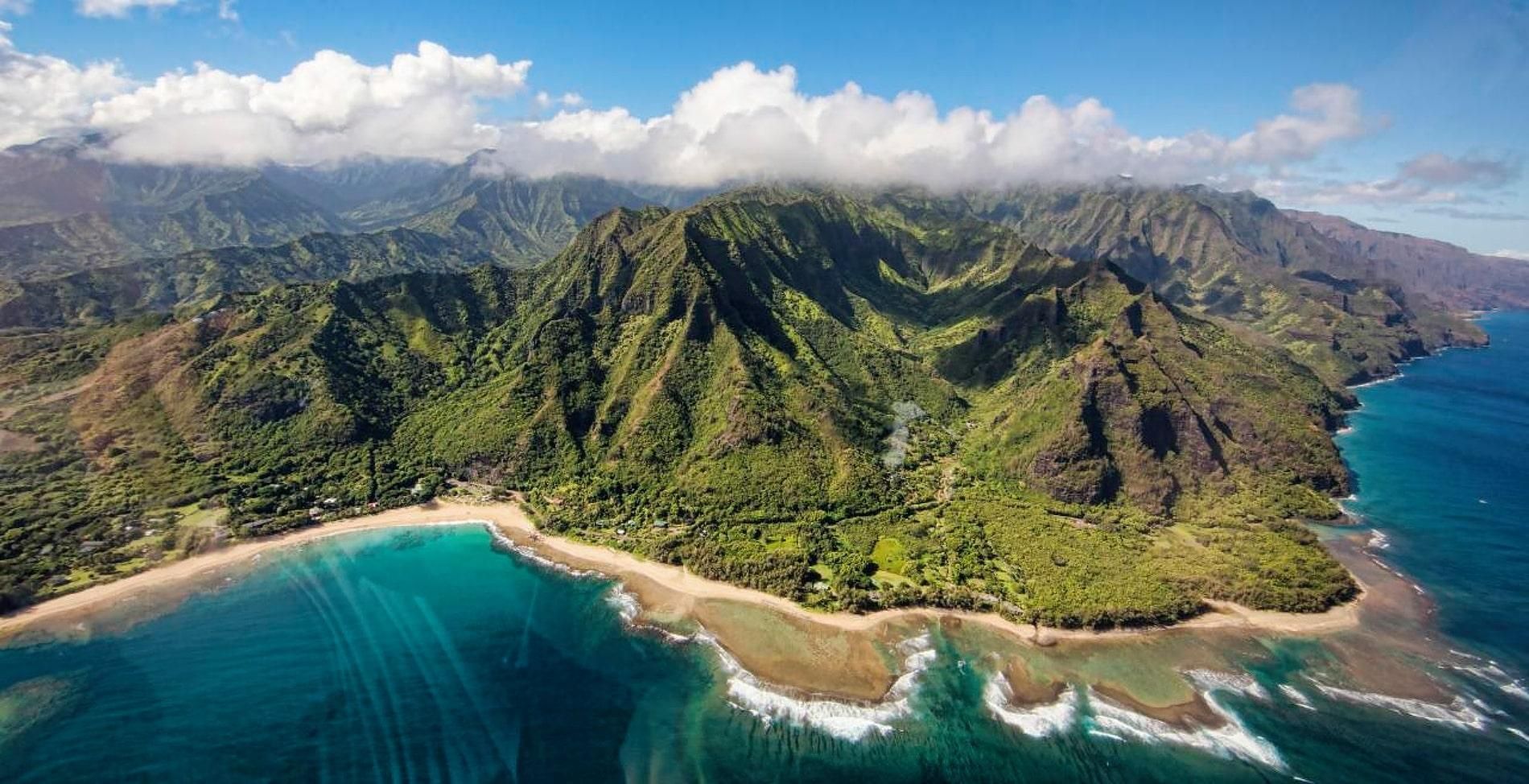 Aerial view of lush green mountains curving around a turquoise tropical bay with sandy beach and waves