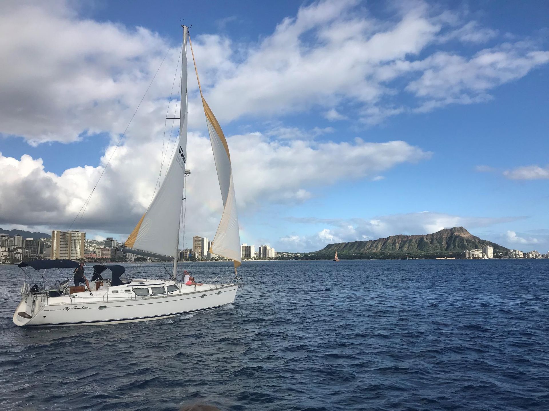 White sailboat on choppy blue water near a city skyline under a bright cloudy sky