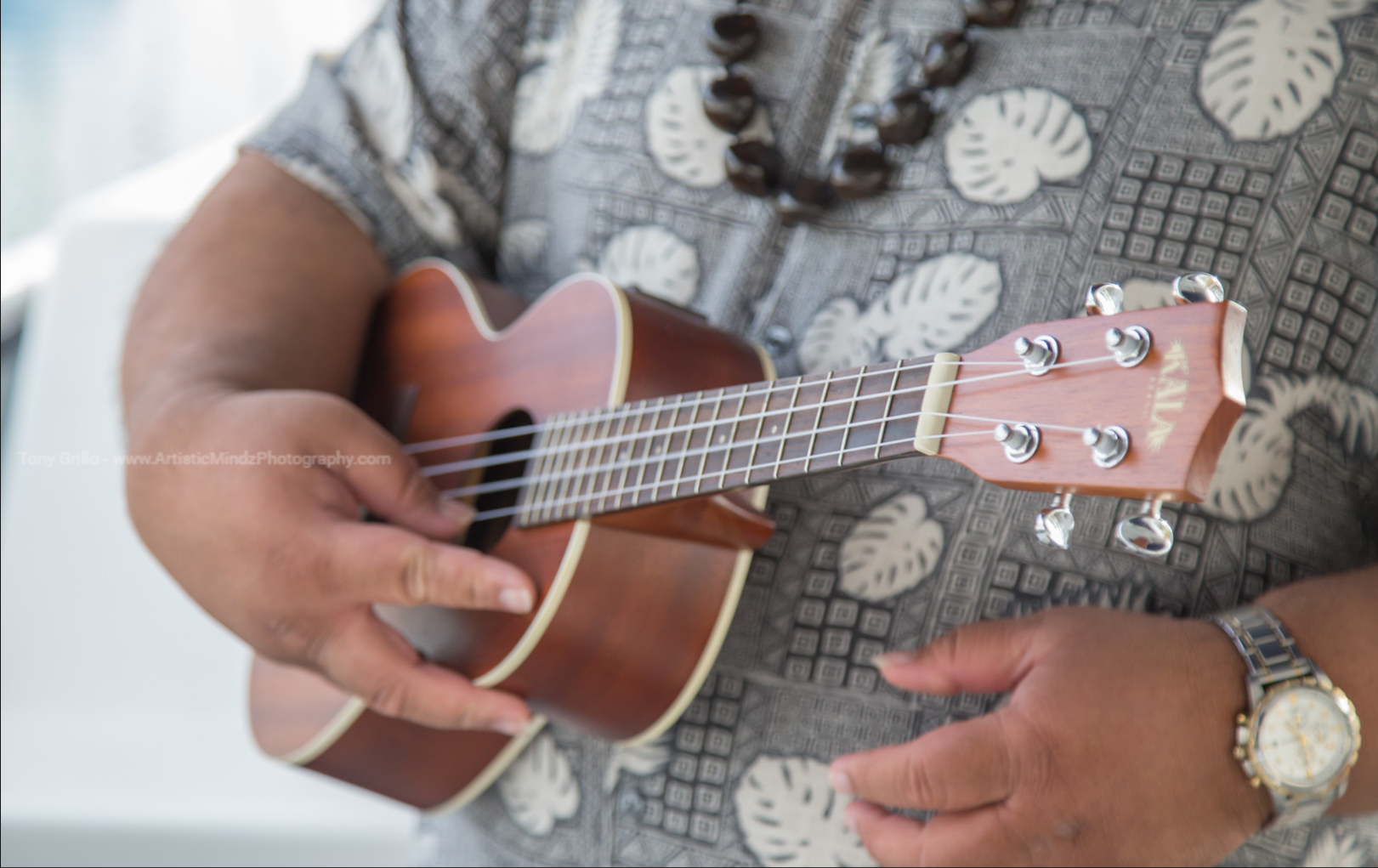 Person playing a small brown ukulele, wearing a patterned shirt and wristwatch.