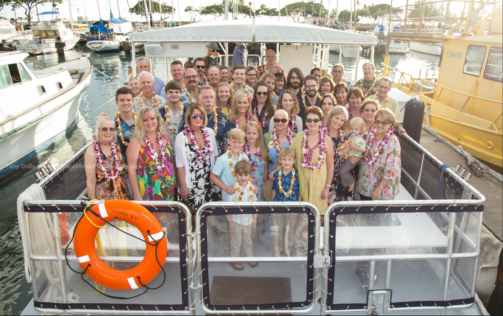 Large group smiling on a boat deck with an orange life ring, docked in a marina.