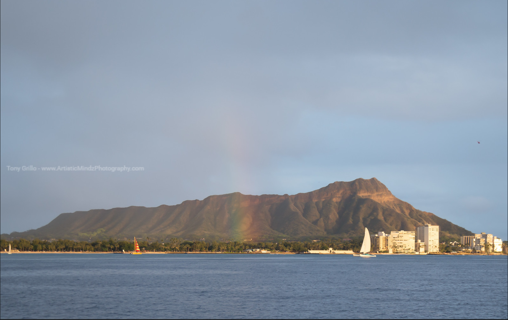 Diamond Head and Waikiki skyline by the ocean under a faint rainbow at sunset