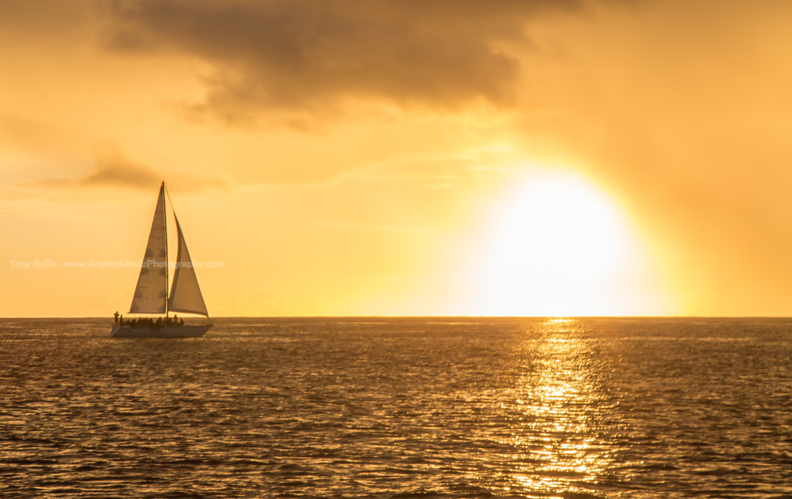 Sailboat on shimmering ocean at sunset with golden sky and bright sun reflection