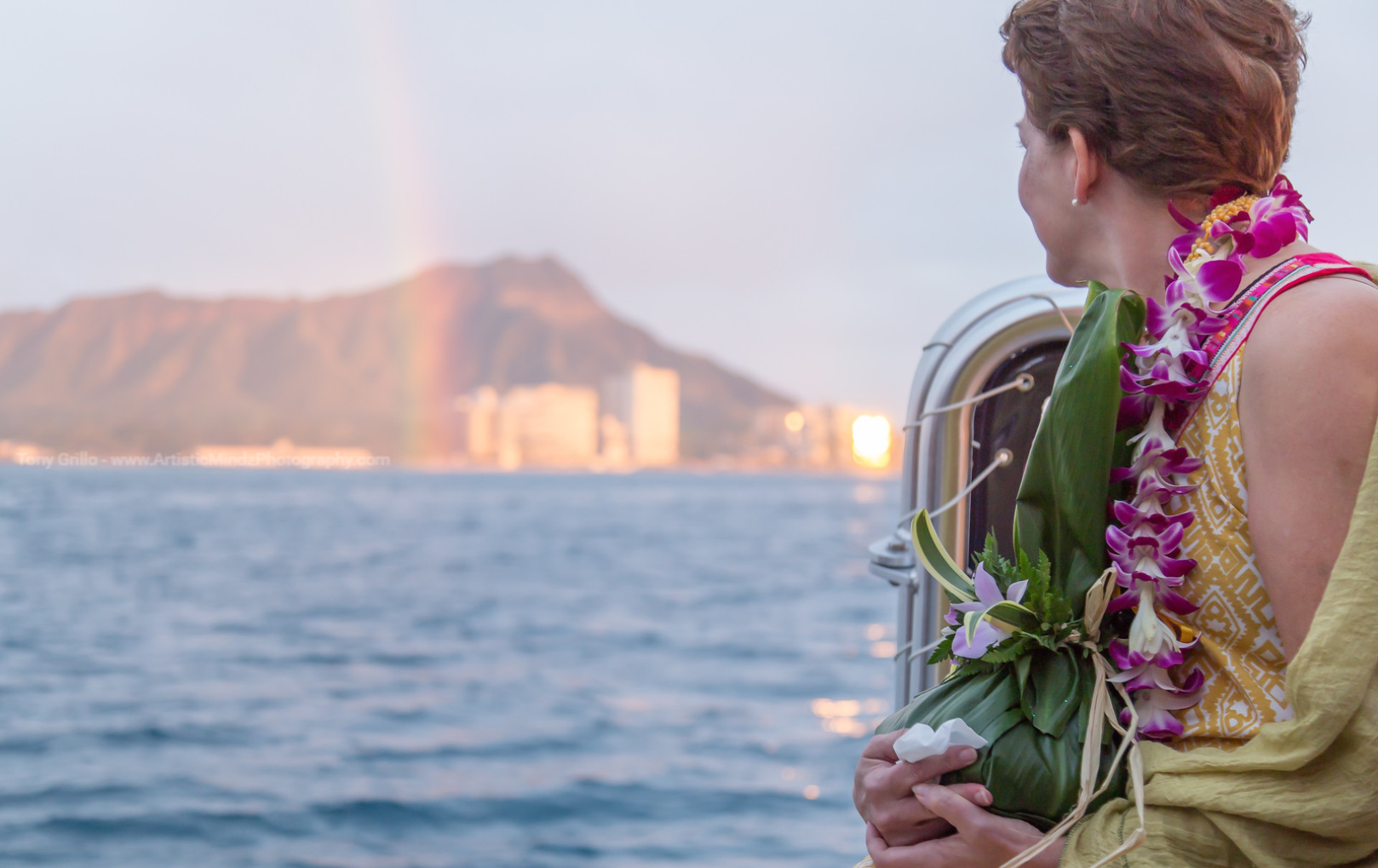 Person on a boat holding a flower lei, looking toward a coastline at sunset.