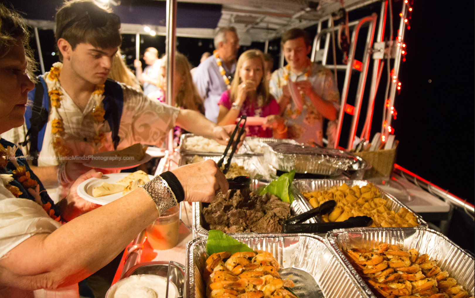 People serving food at a buffet table with trays of rice, vegetables, and skewers at a lively event