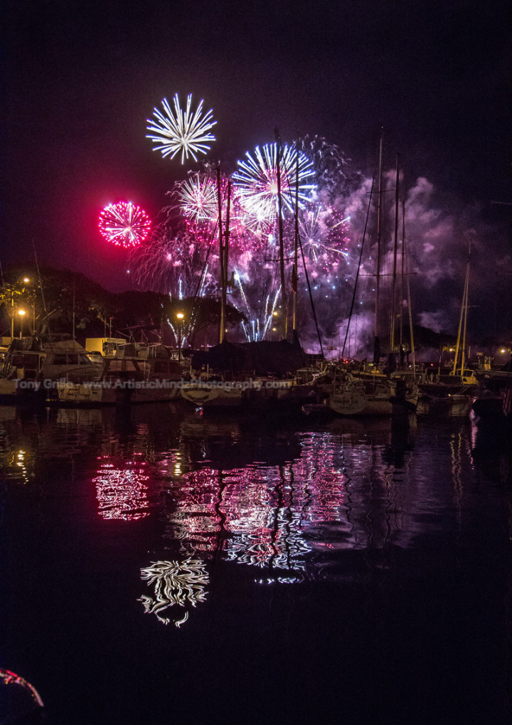 Fireworks bursting over a harbor at night, reflected in the water.
