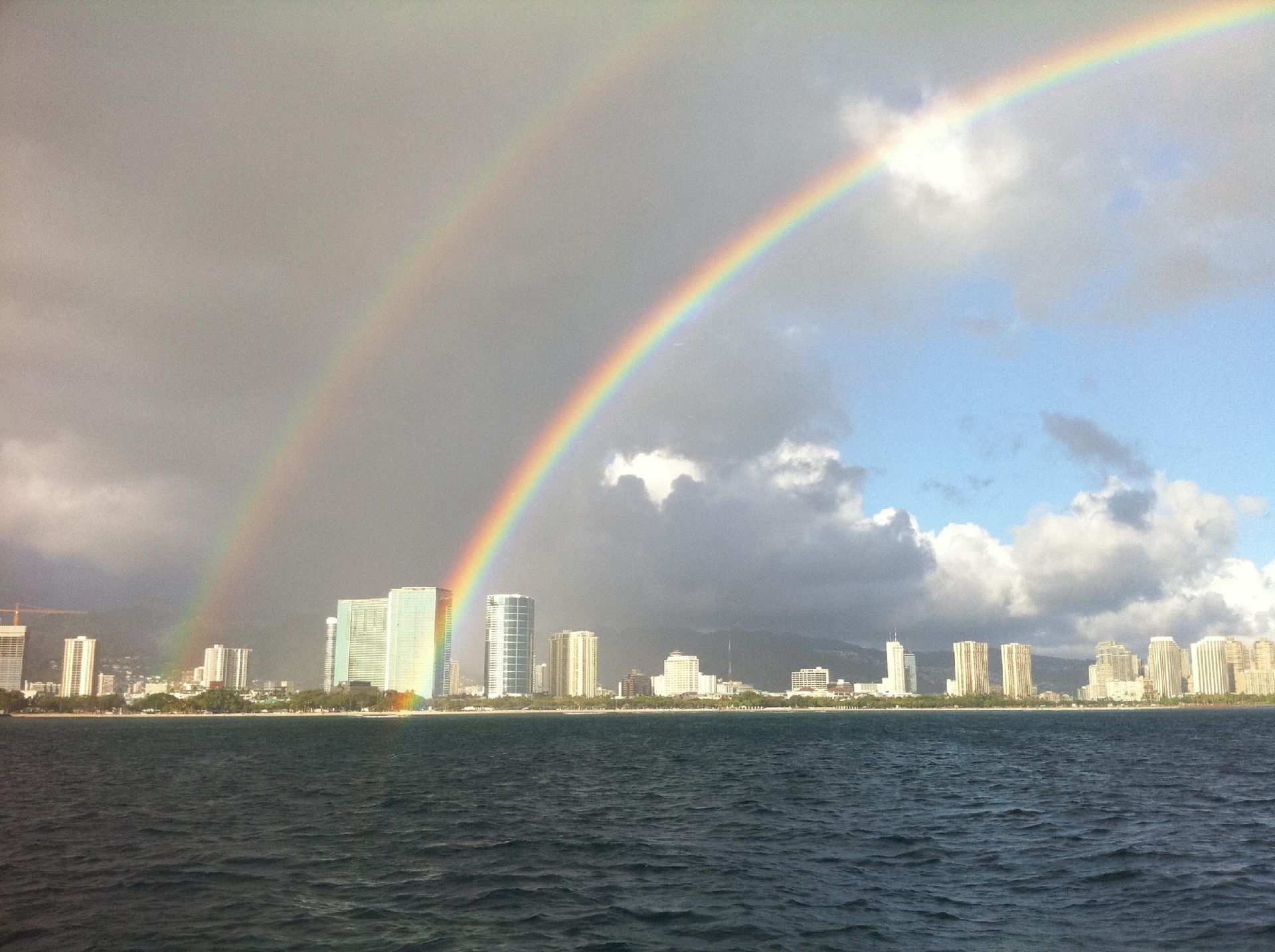 Rainbow over a coastal city skyline above dark water, with clouds and sunlight.