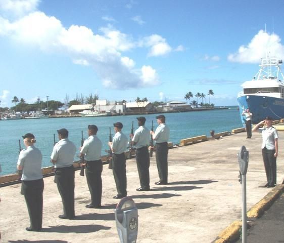 Six uniformed sailors stand at attention on a sunny harbor dock beside a large ship.