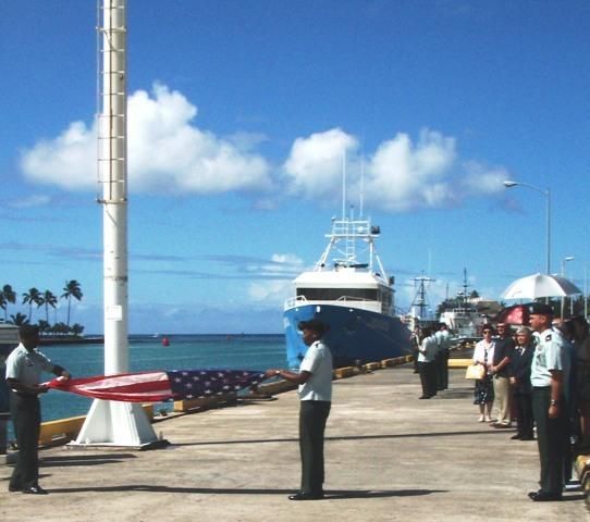 Dockside harbor scene with a large ship, several people, and a red-and-white flag by the water.
