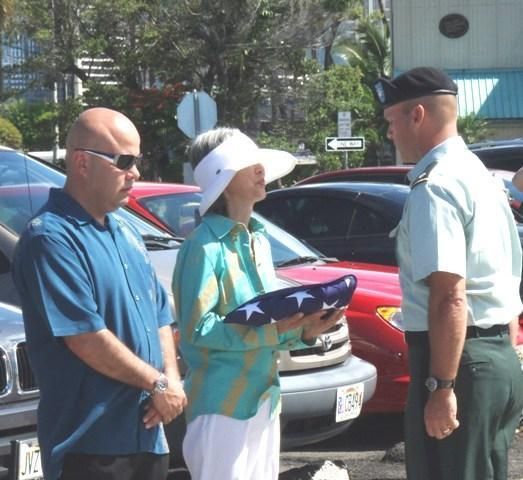 Three people stand by parked cars; one holds a folded umbrella in bright sunlight.