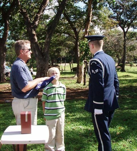 Man handing a paper to a uniformed officer while a child stands nearby in a park