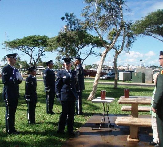 Uniformed officers standing at attention outdoors near a memorial bench and trees under a clear sky