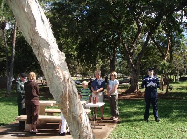 People gathered around picnic tables in a sunny park, with trees and a large white trunk in the foreground.