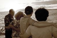 Four friends stand arm in arm, facing a cloudy ocean shoreline at sunset.