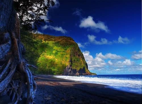 Rocky coastline with a green cliff, dark beach, blue ocean, and cloudy sky
