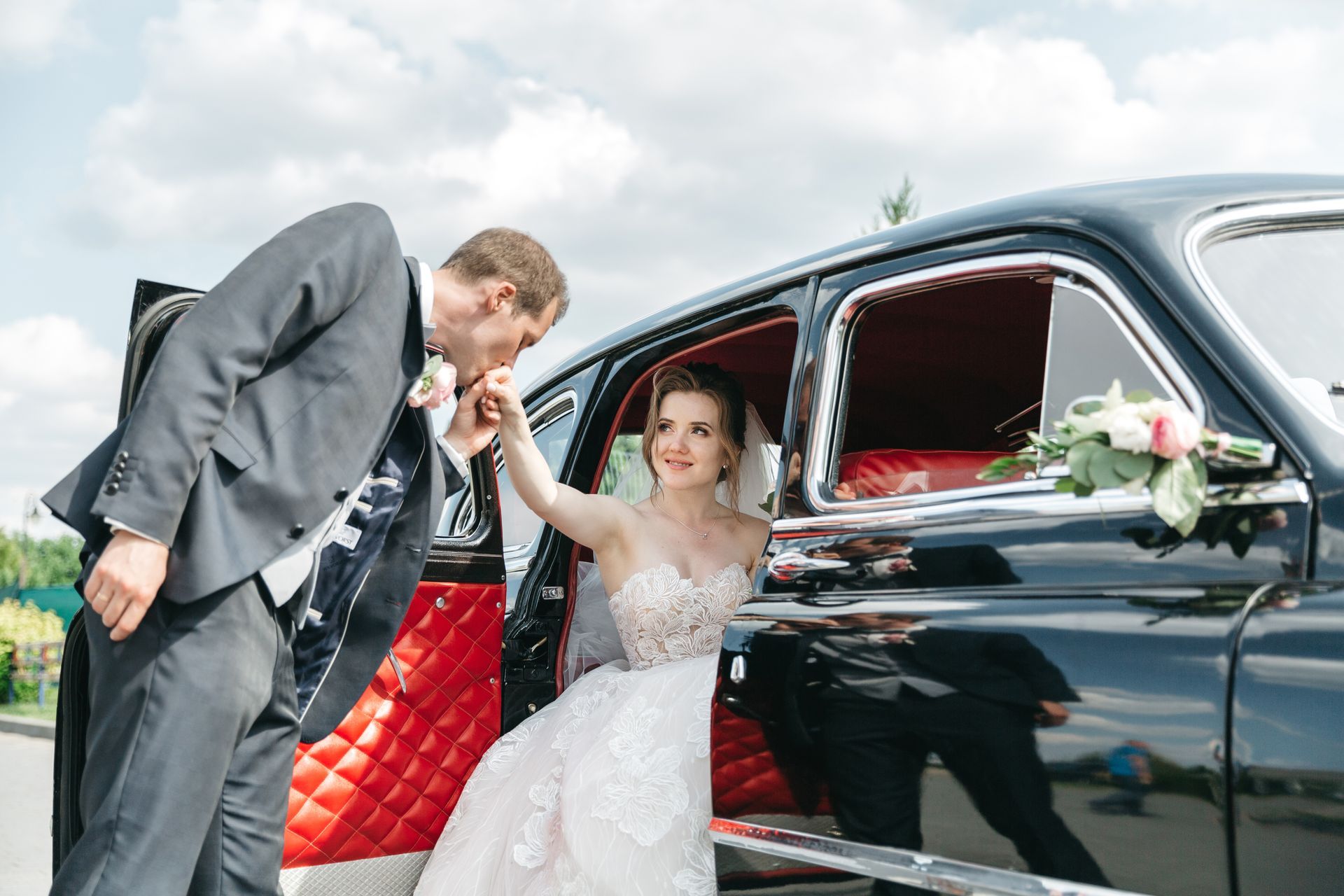 Bride in a white dress kisses a man beside a black vintage car with red interior, bouquet on the seat
