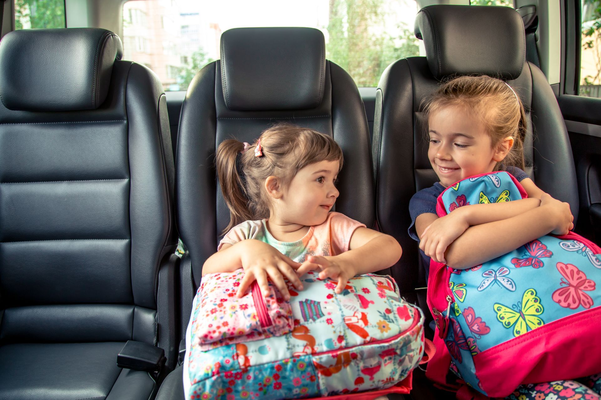 Two children sit in a car’s back seat with colorful backpacks, smiling at each other.