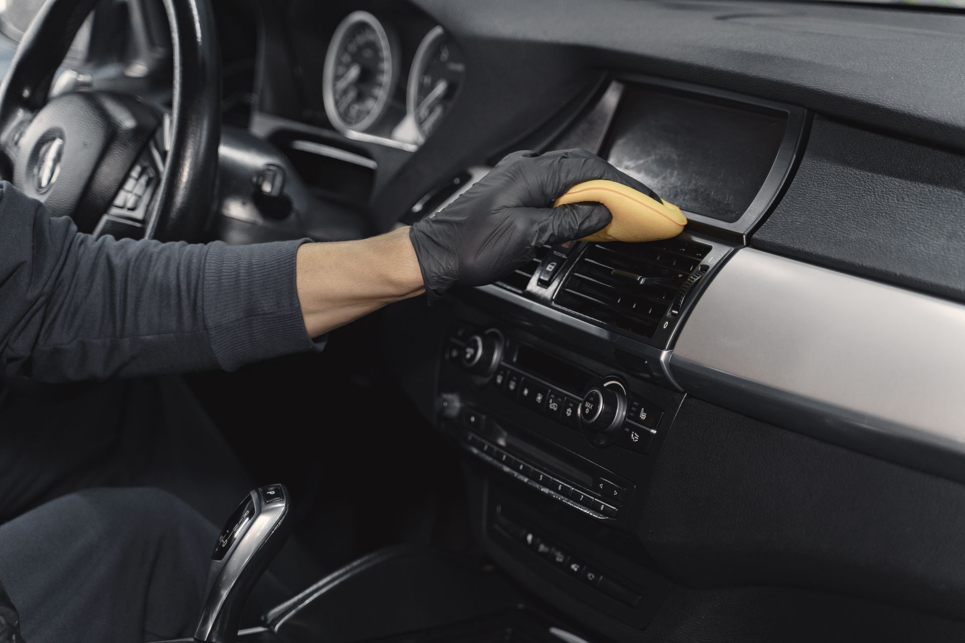 Gloved hand wiping a car dashboard with a yellow cloth inside a luxury vehicle